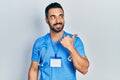 Handsome hispanic man with beard wearing doctor uniform smiling with happy face looking and pointing to the side with thumb up Royalty Free Stock Photo