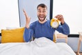 Handsome hispanic man in the bed holding alarm clock celebrating victory with happy smile and winner expression with raised hands Royalty Free Stock Photo