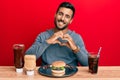 Handsome hispanic man eating a tasty classic burger and soda smiling in love doing heart symbol shape with hands