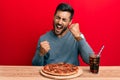 Handsome hispanic man eating tasty pepperoni pizza celebrating surprised and amazed for success with arms raised and eyes closed