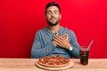 Handsome hispanic man eating tasty pepperoni pizza smiling with hands on chest, eyes closed with grateful gesture on face