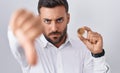 Handsome hispanic man holding cardano cryptocurrency coin with angry face, negative sign showing dislike with thumbs down,