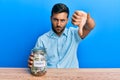 Handsome hispanic man holding charity jar with money with angry face, negative sign showing dislike with thumbs down, rejection Royalty Free Stock Photo