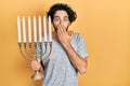 Handsome hispanic man holding menorah hanukkah jewish candle covering mouth with hand, shocked and afraid for mistake Royalty Free Stock Photo