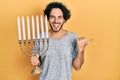 Handsome hispanic man holding menorah hanukkah jewish candle pointing thumb up to the side smiling happy with open mouth Royalty Free Stock Photo