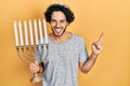 Handsome hispanic man holding menorah hanukkah jewish candle smiling happy pointing with hand and finger to the side Royalty Free Stock Photo