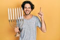 Handsome hispanic man holding menorah hanukkah jewish candle smiling with an idea or question pointing finger with happy face, Royalty Free Stock Photo