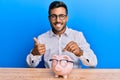 Handsome hispanic man holding piggy bank with glasses smiling happy and positive, thumb up doing excellent and approval sign Royalty Free Stock Photo