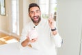 Handsome hispanic man holding piggy bank surprised with an idea or question pointing finger with happy face, number one Royalty Free Stock Photo