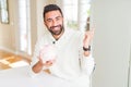 Handsome hispanic man holding piggy bank very happy pointing with hand and finger to the side Royalty Free Stock Photo