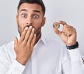 Handsome hispanic man holding polkadot cryptocurrency coin covering mouth with hand, shocked and afraid for mistake