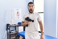 Handsome hispanic man holding therapy massage gun at physiotherapy center scared and amazed with open mouth for surprise, Royalty Free Stock Photo