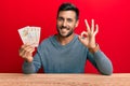 Handsome hispanic man holding united kingdom pounds doing ok sign with fingers, smiling friendly gesturing excellent symbol