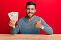 Handsome hispanic man holding united kingdom pounds smiling happy and positive, thumb up doing excellent and approval sign