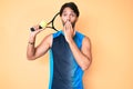 Handsome hispanic man playing tennis holding racket and ball covering mouth with hand, shocked and afraid for mistake