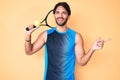 Handsome hispanic man playing tennis holding racket and ball smiling happy pointing with hand and finger to the side Royalty Free Stock Photo