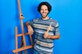 Handsome hispanic man standing by empty easel stand holding brushes sticking tongue out happy with funny expression