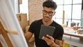 Handsome hispanic man using tablet in art studio with easel and paints Royalty Free Stock Photo