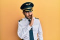 Handsome hispanic man wearing airplane pilot uniform hand on mouth telling secret rumor, whispering malicious talk conversation