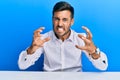 Handsome hispanic man wearing business clothes sitting on the table shouting frustrated with rage, hands trying to strangle, Royalty Free Stock Photo