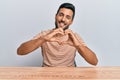 Handsome hispanic man wearing casual clothes sitting on the table smiling in love doing heart symbol shape with hands