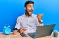 Handsome hispanic man working using laptop at the office pointing thumb up to the side smiling happy with open mouth Royalty Free Stock Photo
