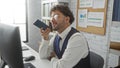 Handsome hispanic man in workplace talking on smartphone at desk with computer and documents in modern office environment Royalty Free Stock Photo