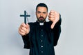 Handsome hispanic priest man with beard holding catholic cross with angry face, negative sign showing dislike with thumbs down,