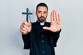 Handsome hispanic priest man with beard holding catholic cross with open hand doing stop sign with serious and confident