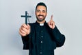 Handsome hispanic priest man with beard holding catholic cross surprised with an idea or question pointing finger with happy face,