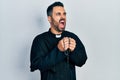 Handsome hispanic priest man with beard praying holding catholic rosary angry and mad screaming frustrated and furious, shouting