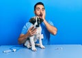 Handsome hispanic veterinary man with beard checking dog health afraid and shocked, surprise and amazed expression with hands on Royalty Free Stock Photo