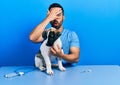 Handsome hispanic veterinary man with beard checking dog health peeking in shock covering face and eyes with hand, looking through Royalty Free Stock Photo