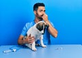 Handsome hispanic veterinary man with beard checking dog health smelling something stinky and disgusting, intolerable smell, Royalty Free Stock Photo