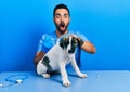 Handsome hispanic veterinary man with beard putting vaccine to puppy dog afraid and shocked with surprise and amazed expression, Royalty Free Stock Photo