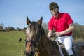 Handsome Male Horse Rider on horseback with white breeches, black boots and red polo shirt in green field with horses in distance Royalty Free Stock Photo