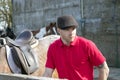 Handsome horse rider in red polo shirt and flatcap standing next to his saddled horse