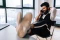 Handsome Indian business man in formalwear talking on mobile phone sitting with feet on table with laptop in office by Royalty Free Stock Photo