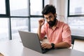 Handsome Indian businessman in casual clothes sitting at desk with laptop looking at camera holding glasses in light Royalty Free Stock Photo