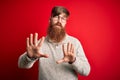 Handsome Irish redhead man with beard wearing casual sweater and glasses over red background Moving away hands palms showing Royalty Free Stock Photo