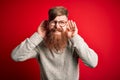 Handsome Irish redhead man with beard wearing casual sweater and glasses over red background Trying to hear both hands on ear Royalty Free Stock Photo