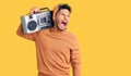 Handsome latin american young man holding boombox, listening to music angry and mad screaming frustrated and furious, shouting Royalty Free Stock Photo