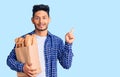 Handsome latin american young man holding paper bag with bread with a big smile on face, pointing with hand finger to the side Royalty Free Stock Photo