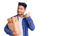 Handsome latin american young man holding paper bag with bread smiling doing phone gesture with hand and fingers like talking on Royalty Free Stock Photo