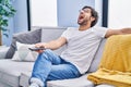 Handsome latin man holding television remote control angry and mad screaming frustrated and furious, shouting with anger looking Royalty Free Stock Photo