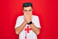 Handsome lifeguard man wearing t-shirt with red cross and whistle over isolated background shocked covering mouth with hands for