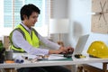 Male construction engineer in white shirt and yellow vest using laptop computer and working with blueprints. Royalty Free Stock Photo