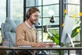 Handsome male programmer sits in a headset in the office at the table in profile, works at the computer, smiles and