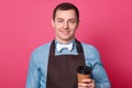 Handsome male waiter holds takeaway coffee wears shirt with bowtie, apron, holds paper cup of drink, own coffee shop, has gentle Royalty Free Stock Photo