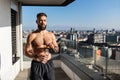 Handsome man with bare chest standing on balcony and enjoying beautiful view on city. Concept of selfcare and enjoying